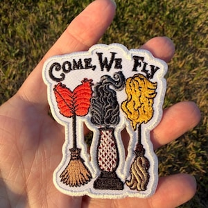 May include: An embroidered patch with a white background featuring the black text "COME, WE FLY" above three distinct items: a red broom, a black vacuum cleaner with a patterned base, and a yellow mop, all with detailed stitching.