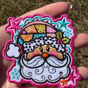 May include: An embroidered patch featuring a Santa Claus face with a white beard, a colorful patchwork hat, and holly berries. The design is accented with teal snowflakes and pink stars, all outlined with a vibrant pink scalloped border.
