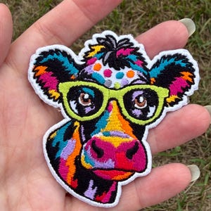 May include: An embroidered patch featuring a cow head with a multi-colored pattern of bright pink, orange, yellow, blue, and black. The cow wears lime green glasses with black lenses and has a white border around the entire shape.