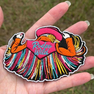May include: An embroidered patch featuring a figure wearing a pink cowboy hat and an orange jacket with a pink bandana that reads Rodeo Wild in blue script. The design includes long, multicolored fringe in yellow, blue, orange, and white.