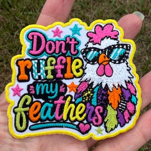 May include: An embroidered patch featuring a white chicken wearing teal and black sunglasses. The text reads Don't ruffle my feathers in colorful, bold lettering. The design includes small stars and a heart, all outlined in a bright yellow border.