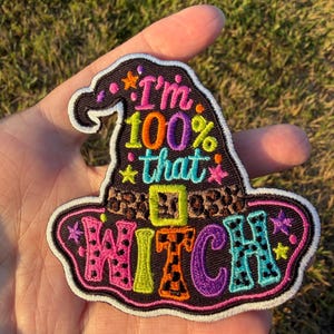May include: A black embroidered patch shaped like a witch hat with a leopard print band. The text reads I'm 100% that WITCH in colorful, patterned lettering. The design is accented with small embroidered stars and dots in pink, orange, and yellow.