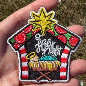 May include: An embroidered patch shaped like a stable with a yellow star on top. The black center features the white text O Holy Night above a manger with a figure. The roof is green with red circles, and the side pillars have red and white stripes.