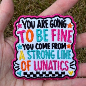 May include: A square embroidered patch with a scalloped hot pink border on a white background. The text reads: YOU ARE GOING TO BE FINE YOU COME FROM A STRONG LINE OF LUNATICS. The lettering is in black, hot pink, orange, teal, and yellow.
