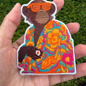 May include: An embroidered patch featuring a brown monkey wearing bright orange sunglasses and a vibrant shirt with a colorful floral pattern in shades of pink, orange, yellow, and turquoise, finished with a white border edge.