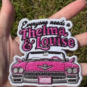 May include: A white embroidered patch featuring a pink vintage convertible car at the bottom and the black and pink text Everyone needs a Thelma & Louise at the top. The patch has a white border and detailed stitching of the car's front grille.