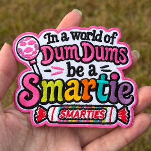 May include: An embroidered patch with a white background featuring a pink lollipop and the text In a world of Dum Dums be a Smartie. The word Smartie is in multi-colored letters, and a Smarties candy roll is depicted at the bottom in red and white.