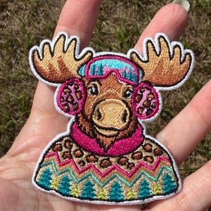 May include: An embroidered patch featuring a brown moose wearing a bright pink scarf, leopard print earmuffs, and blue ski goggles. The moose is dressed in a patterned sweater with a colorful geometric design and small green pine trees.