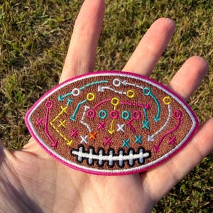Può includere: Una toppa ricamata di forma ovale che raffigura un pallone da football americano, con uno sfondo marrone testurizzato e un dettaglio di lacci bianchi cuciti sul fondo. La superficie presenta schemi di gioco colorati con linee, frecce e croci.