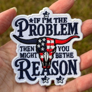 May include: A white embroidered patch with a dark blue border featuring the text IF I'M THE PROBLEM THEN YOU MIGHT BE THE REASON. In the center is a steer skull graphic colored with the American flag pattern of red, white, and blue stars and stripes.