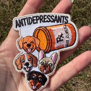 May include: An embroidered patch featuring an orange prescription bottle with the text ANTIDEPRESSANTS above it. Below the bottle are four dog faces: a golden retriever, a beagle, a French bulldog, and a Rottweiler, all smiling.