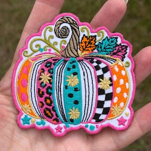 May include: An embroidered patch shaped like a pumpkin with a bright pink border. The pumpkin features vertical segments with varied patterns including leopard print, black and white checks, polka dots, and stripes, accented with small gold star embroidery.