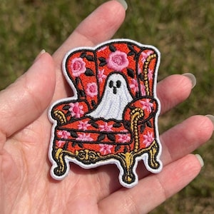 May include: An embroidered patch featuring a white ghost sitting in a red armchair with a floral pattern. The chair has gold-colored legs and armrests, and the entire patch is outlined in white thread against a dark background.