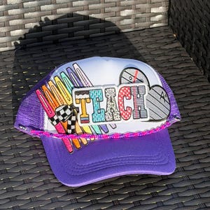 May include: A purple trucker hat with a white front panel and a purple mesh back. The hat features the word "TEACH" in colourful, textured letters. Additional patches include a chequered flag and a shield design.