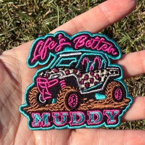 May include: An embroidered patch featuring an off-road vehicle with a leopard print pattern, pink accents, and brown mud details. The text Life's Better is stitched in pink above the vehicle, and the word MUDDY is stitched in pink and teal below.