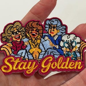May include: An embroidered patch featuring four cartoon characters sitting on a couch with the text Stay Golden in yellow script. The characters have distinct hair colors including blonde, brown, grey, and white, and wear colorful clothing.
