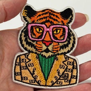 May include: An embroidered patch featuring a tiger wearing pink-rimmed glasses, a green shirt, and a tan blazer with a repeating dark brown pattern. The tiger has orange fur with black stripes and a gold chain necklace, all outlined in white thread.