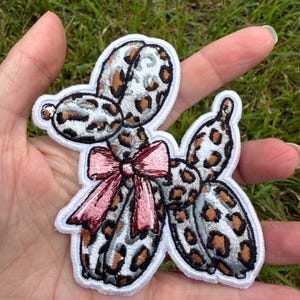 May include: An embroidered patch shaped like a balloon animal dog with a leopard print pattern in shades of brown, black, and grey. The dog figure features a prominent light pink bow tied around its neck and is outlined with a white border.