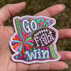 May include: An embroidered patch featuring a colorful cheerleading pom-pom, a football with a leopard print pattern, and a pink star. The text reads GO FIGHT WIN in white and purple lettering against a bright, multi-colored background.