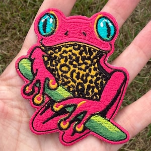May include: An embroidered patch featuring a bright pink frog with large turquoise eyes and a yellow chest patterned with black leopard spots. The frog is depicted resting on a green branch, with its limbs outlined in black and yellow.