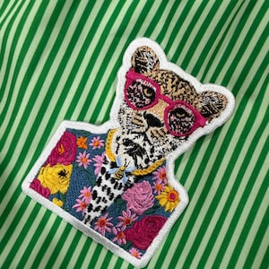 May include: Embroidered patch of a leopard wearing pink sunglasses and a gold chain. The leopard is set against a floral and denim background, with a white border. The patch is on a green and white striped fabric.