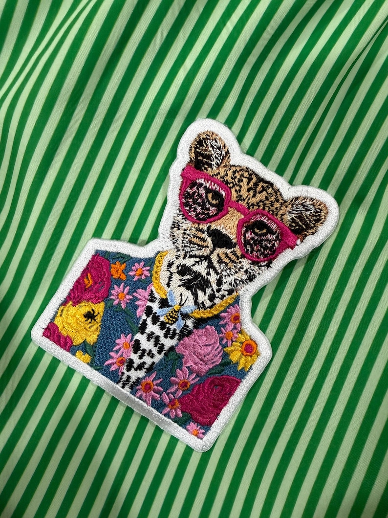 May include: Embroidered patch of a leopard wearing pink sunglasses and a gold chain. The leopard is set against a floral and denim background, with a white border. The patch is on a green and white striped fabric.
