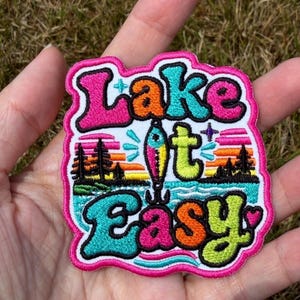 May include: An embroidered patch with a bright pink border featuring the text Lake it Easy in colourful bubble letters. The design includes a fishing lure, a sunset over a lake, pine trees, and small star accents in shades of blue, orange, and green.
