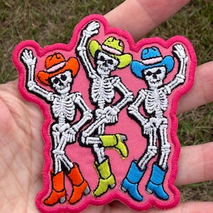 May include: An embroidered patch featuring three white skeletons dancing on a bright pink background. Each skeleton wears a colorful cowboy hat and matching cowboy boots in shades of orange, lime green, and blue with pink accents.