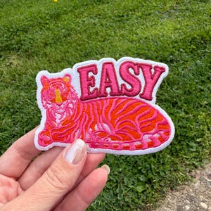 Puede incluir: Parche bordado con un tigre estilizado en tonos rosa y naranja, con la palabra "EASY" en letras rosas. El parche tiene un borde blanco. El parche se sostiene sobre un fondo de hierba verde.