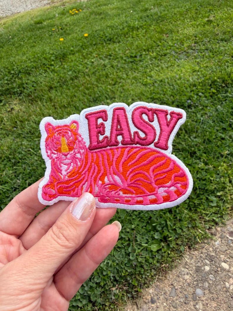 May include: Embroidered patch featuring a stylized tiger in shades of pink and orange, with the word "EASY" in pink lettering. The patch has a white border and is held against a background of green grass.