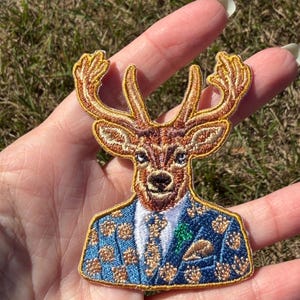 May include: An embroidered patch featuring a deer head with large antlers, wearing a blue suit jacket with a gold floral pattern, a white shirt, and a matching gold floral tie, with a small green accent on the lapel.
