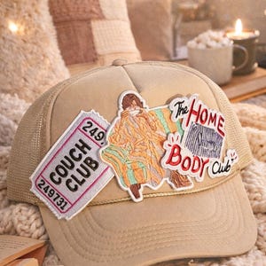 Pode incluir: Boné de caminhoneiro bege com costas em malha, com patches bordados. Os patches incluem um bilhete que diz "COUCH CLUB" e uma pessoa em um sofá com as palavras "The HOME BODY Club".