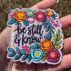 May include: A rectangular embroidered patch with a light purple background featuring the black text "be still & know" surrounded by a vibrant floral border of orange, magenta, and teal flowers with green leaves and small yellow dots.