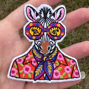 May include: An embroidered patch featuring a zebra wearing large, colorful, patterned glasses and a blue neckerchief with yellow accents. The zebra is dressed in a bright red jacket adorned with a repeating pink and green floral pattern.