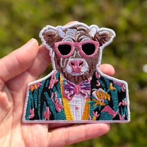 May include: An embroidered patch featuring a brown cow wearing pink-rimmed sunglasses and a patterned bow tie. The cow is dressed in a teal and black jacket with a yellow vest and a floral lapel detail, all outlined in a white stitched border.