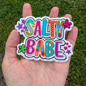 May include: An embroidered patch featuring the text SALTY BABE in colorful, multi-patterned letters with a white border. The design includes small purple stars, a pink flower, a green flower, and three small pink hearts on the edges.