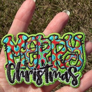 May include: An embroidered patch featuring the word MERRY in a bubble-style font with a red, lime green, and bright blue polka dot pattern, and the word Christmas in a black cursive script font, all outlined in a vibrant lime green border.