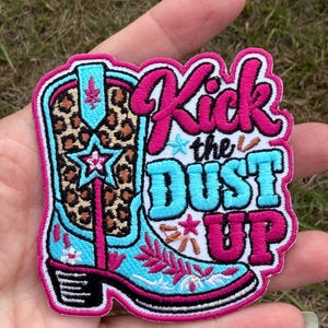 May include: An embroidered patch featuring a bright blue cowboy boot with a leopard print shaft, a pink star, and floral accents. The text reads Kick the DUST UP in bold pink and blue lettering, accompanied by small star and boot icons.