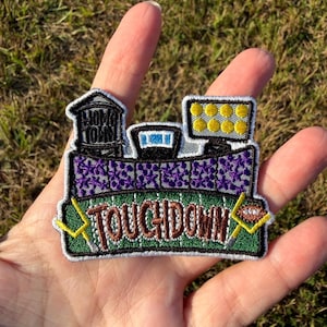 Op de afbeelding: Een geborduurde patch met een footballstadion-scène, een watertoren met het label HOME TOWN, een scorebord en stadionverlichting. Het groene veld toont het woord TOUCHDOWN in bruine letters, geflankeerd door twee gele doelpalen.