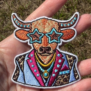 May include: An embroidered patch featuring a bull wearing star-shaped glasses, a light blue blazer with leopard print lapels, and a pink trim. The bull has orange fur, turquoise accents on its horns, and a layered necklace with a circular pendant.