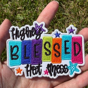 May include: A rectangular embroidered patch featuring the text Highly Blessed Hot Mess in black script and block lettering. The design includes colorful vertical bars in teal, blue, purple, lime, orange, and pink, accented with small stars.