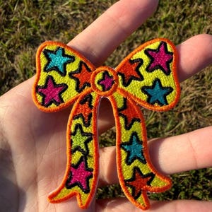May include: An embroidered bow-shaped patch featuring a bright yellow base with a repeating pattern of stars in vibrant shades of hot pink, turquoise, and orange. The entire design is outlined in a bold orange thread border.