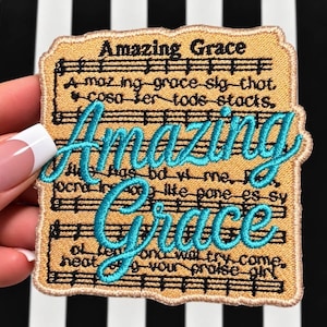 May include: A rectangular embroidered patch with a tan background featuring black musical staff lines and lyrics. The words Amazing Grace are stitched in large, bright turquoise cursive script over the center of the black text.