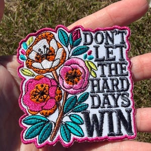May include: A square embroidered patch with a white background featuring a floral arrangement of orange and pink flowers with teal leaves. The text DON'T LET THE HARD DAYS WIN is stitched in black thread on the right side, bordered by a pink edge.