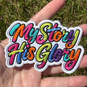 May include: A white embroidered patch featuring the text My Story His Glory in a multi-colored, bubble-style font. The letters are filled with vibrant shades of pink, orange, lime green, blue, and purple, all outlined in a bold black thread.