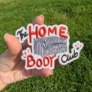 May include: Embroidered patch with the text "The Home Body Club" in red and black. The patch features a stylized couch in blue and white. The patch is white with a black border and is held against a background of green grass.