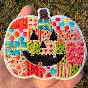 May include: A pumpkin-shaped embroidered patch featuring a patchwork design of various colorful patterns including teal plaid, pink flowers, orange stripes, small red hearts, and polka dots, with a black stitched jack-o'-lantern face.