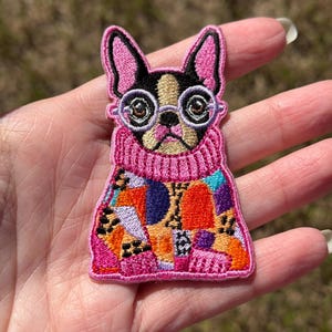 May include: An embroidered patch featuring a French Bulldog wearing round glasses and a pink turtleneck sweater. The dog's body is covered in a colorful, multi-patterned design with shades of orange, purple, blue, and black animal print accents.