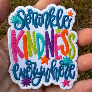 Puede incluir: Un parche bordado blanco con borde festoneado que presenta el texto Sprinkle KINDNESS everywhere en hilo azul, rosa, naranja, verde lima y morado. El diseño incluye pequeñas formas de estrellas bordadas en naranja, turquesa y rosa.