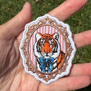 May include: An embroidered patch featuring an orange and white tiger wearing a bright blue bow tie. The tiger is centered within a decorative bronze-colored oval frame against a background of vertical pink and white stripes with a white border.